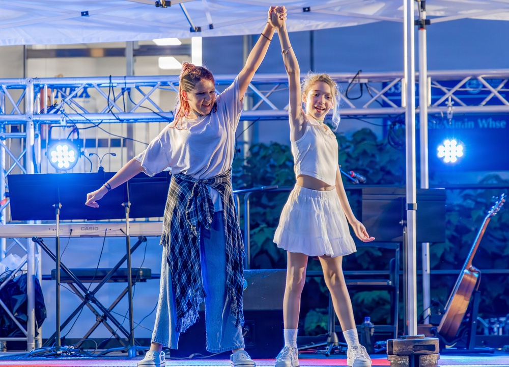 Two young females are performing a dance routine on an outdoor stage, holding raised hands together in a choreographed pose. The stage is equipped with professional lighting rigs, a Casio keyboard, and a guitar visible in the background. Both performers are dressed in white outfits and appear to be smiling during their performance.