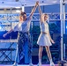 Two young females are performing a dance routine on an outdoor stage, holding raised hands together in a choreographed pose. The stage is equipped with professional lighting rigs, a Casio keyboard, and a guitar visible in the background. Both performers are dressed in white outfits and appear to be smiling during their performance.
