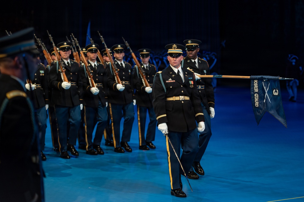 A U.S. Army Honor Guard unit is performing a formal ceremonial drill or parade on an indoor stage illuminated by blue lighting. The lead soldier carries a guidon pennant marked 'HONOR GUARD' with crossed sabers insignia, while the formation behind carries rifles at shoulder arms. The soldiers are dressed in Army Service Uniforms (ASUs) with white gloves, indicating a formal ceremonial event.