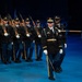 A U.S. Army Honor Guard unit is performing a formal ceremonial drill or parade on an indoor stage illuminated by blue lighting. The lead soldier carries a guidon pennant marked 'HONOR GUARD' with crossed sabers insignia, while the formation behind carries rifles at shoulder arms. The soldiers are dressed in Army Service Uniforms (ASUs) with white gloves, indicating a formal ceremonial event.