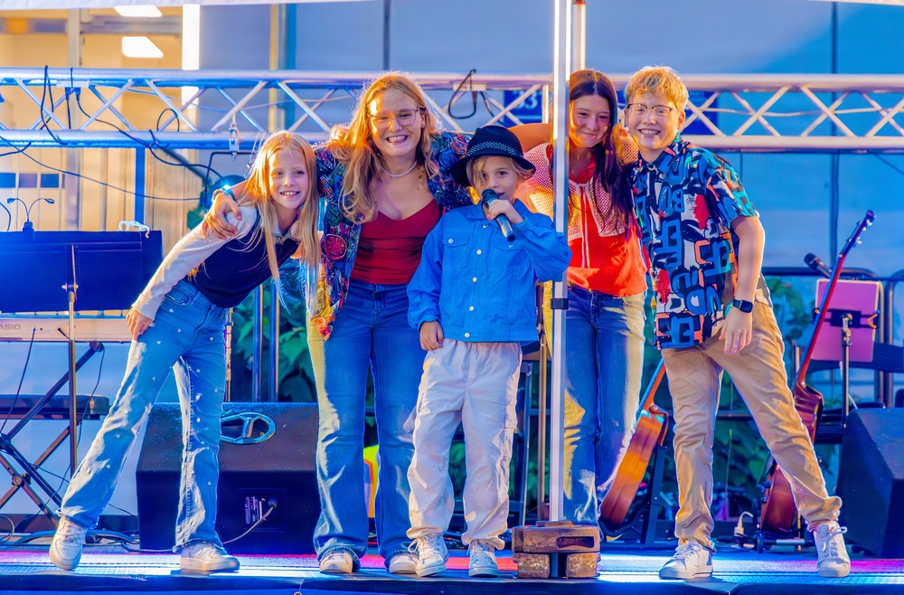 Five young people are posing together on a performance stage, smiling at the camera. The central figure, a small child wearing a black bucket hat and blue denim jacket, is holding a microphone, suggesting a live performance or talent show event. The group appears to be in a celebratory, post-performance moment under stage lighting.