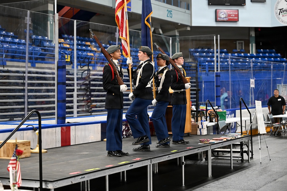 A military color guard, likely JROTC cadets, performs a flag ceremony on a portable stage inside what appears to be an ice hockey arena. Four uniformed cadets are present — two carrying flags (American flag and a state/unit flag) and two carrying ceremonial rifles. The arena seating is largely empty, suggesting this is a pre-event or ceremony setup.