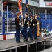 A military color guard, likely JROTC cadets, performs a flag ceremony on a portable stage inside what appears to be an ice hockey arena. Four uniformed cadets are present — two carrying flags (American flag and a state/unit flag) and two carrying ceremonial rifles. The arena seating is largely empty, suggesting this is a pre-event or ceremony setup.