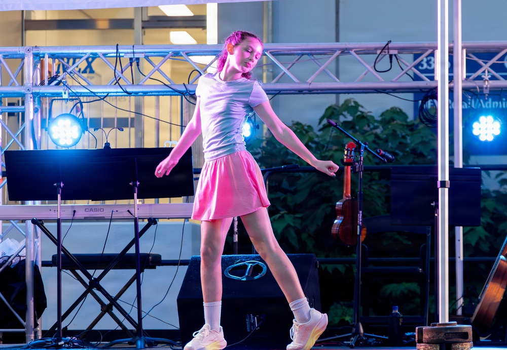 A young girl is performing on a stage, dancing or balancing under pink and blue stage lighting. The stage is equipped with musical instruments and equipment, including a Casio keyboard and a ukulele or small guitar on a stand. The setting appears to be an outdoor or semi-outdoor performance venue.