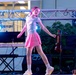 A young girl is performing on a stage, dancing or balancing under pink and blue stage lighting. The stage is equipped with musical instruments and equipment, including a Casio keyboard and a ukulele or small guitar on a stand. The setting appears to be an outdoor or semi-outdoor performance venue.