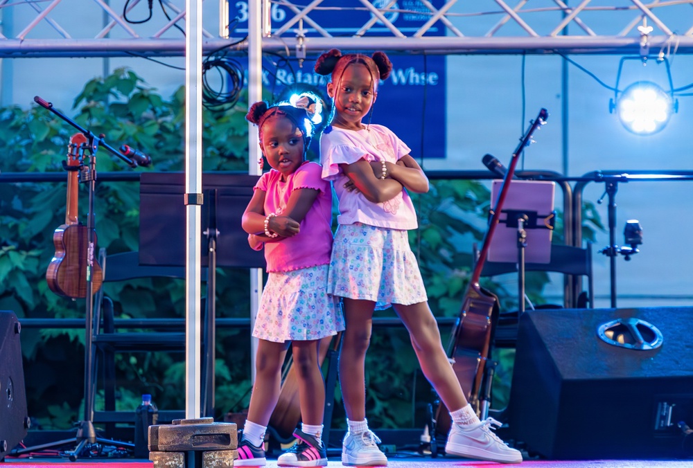 Two young girls are posing confidently on a performance stage with their arms crossed, appearing to be part of a dance or talent show. They are dressed in matching outfits consisting of white floral skirts and colored tops. The stage is equipped with musical instruments, microphone stands, speakers, and stage lighting.