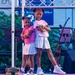 Two young girls are posing confidently on a performance stage with their arms crossed, appearing to be part of a dance or talent show. They are dressed in matching outfits consisting of white floral skirts and colored tops. The stage is equipped with musical instruments, microphone stands, speakers, and stage lighting.
