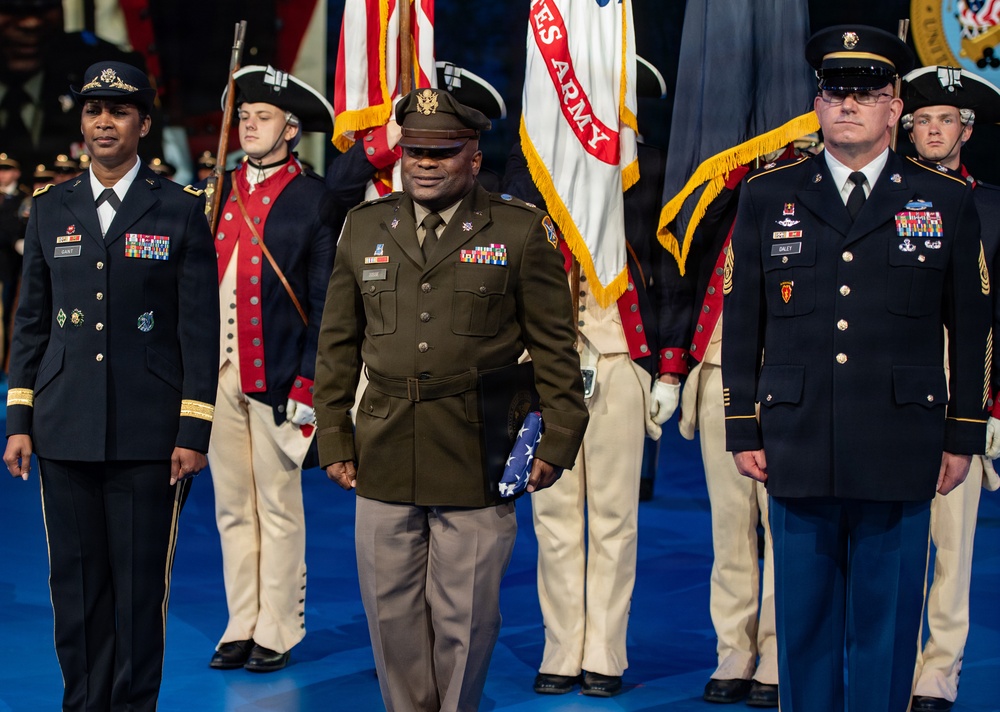 A formal U.S. Army military ceremony featuring personnel in various dress uniforms standing on a blue floor. Three prominent figures are in the foreground — a female officer in Army Service Uniform (blue), a senior officer in the historic Army Green Service Uniform holding a folded American flag, and a male senior NCO in Army Service Uniform (blue). Behind them, members of a ceremonial color guard in Revolutionary War-era uniforms carry flags including a U.S. Army flag.