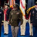 A formal U.S. Army military ceremony featuring personnel in various dress uniforms standing on a blue floor. Three prominent figures are in the foreground — a female officer in Army Service Uniform (blue), a senior officer in the historic Army Green Service Uniform holding a folded American flag, and a male senior NCO in Army Service Uniform (blue). Behind them, members of a ceremonial color guard in Revolutionary War-era uniforms carry flags including a U.S. Army flag.