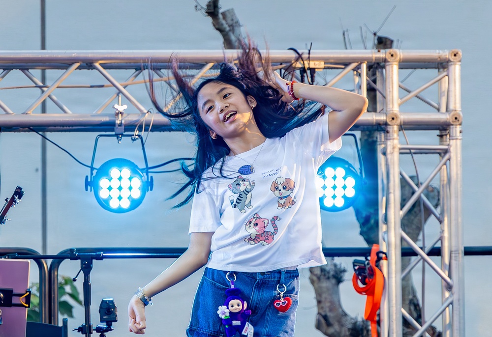 A young girl is performing energetically on an outdoor stage, tossing her long dark hair with a joyful expression. She is wearing a white graphic t-shirt featuring cartoon animals and denim shorts with small charm accessories. The stage is equipped with blue LED stage lights mounted on metal truss structures.