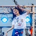 A young girl is performing energetically on an outdoor stage, tossing her long dark hair with a joyful expression. She is wearing a white graphic t-shirt featuring cartoon animals and denim shorts with small charm accessories. The stage is equipped with blue LED stage lights mounted on metal truss structures.