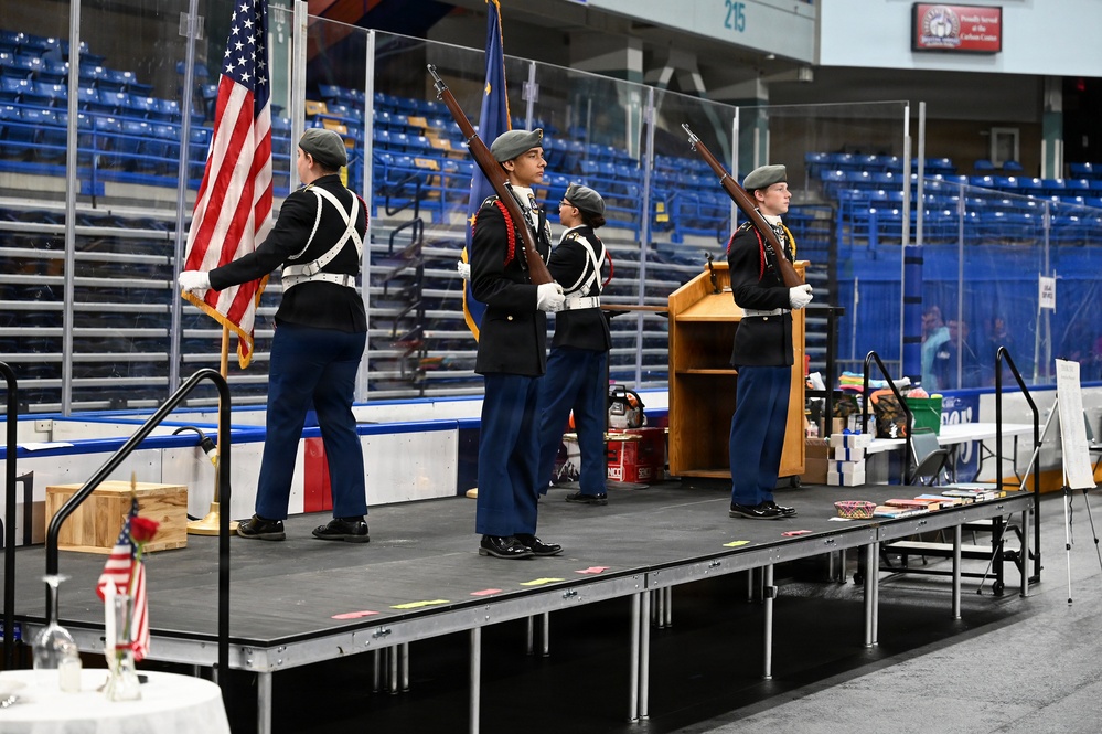 A military honor guard of four cadets in dress uniforms performs a color guard ceremony on a portable stage inside what appears to be an ice hockey arena. The cadets are carrying the American flag and rifles, positioned in formal parade formation. The venue appears to be set up for a special event, with tables and items visible in the foreground.