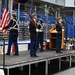 A military honor guard of four cadets in dress uniforms performs a color guard ceremony on a portable stage inside what appears to be an ice hockey arena. The cadets are carrying the American flag and rifles, positioned in formal parade formation. The venue appears to be set up for a special event, with tables and items visible in the foreground.