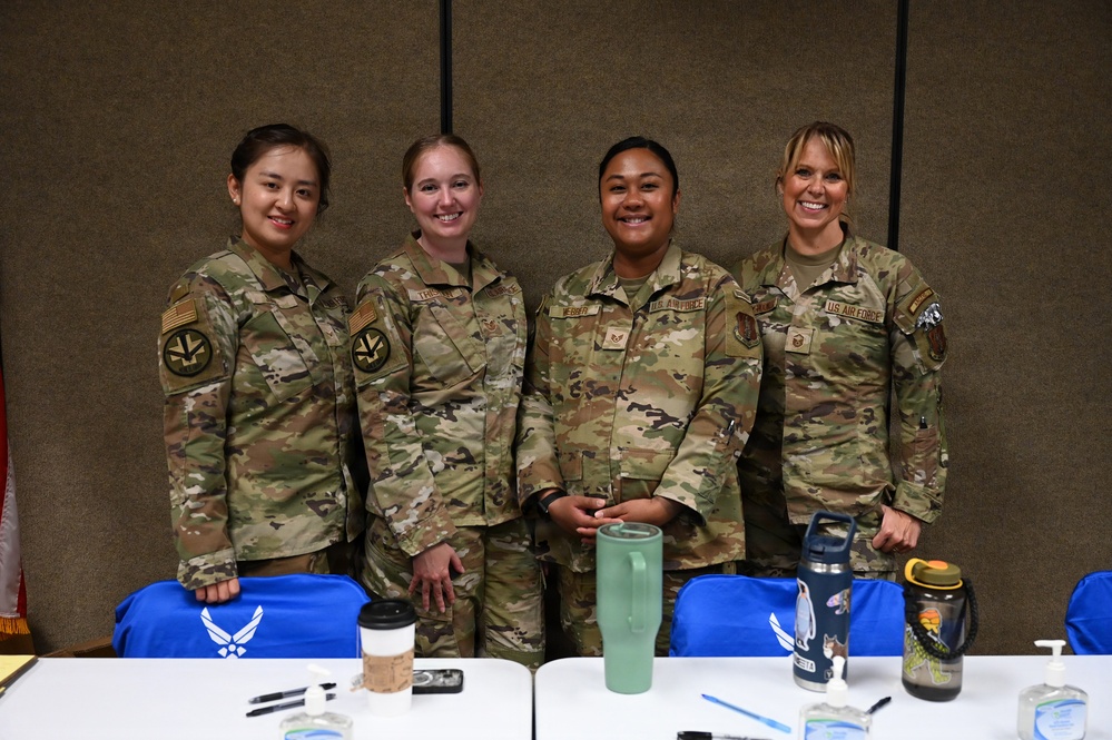 Four female U.S. Air Force personnel in OCP (Operational Camouflage Pattern) uniforms pose together smiling in what appears to be a conference or meeting room. Blue Air Force-branded t-shirts with the Air Force symbol are visible on the table in front of them, suggesting a recruitment, outreach, or special event setting. The group appears to be in good spirits during what seems to be an official military function.