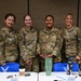 Four female U.S. Air Force personnel in OCP (Operational Camouflage Pattern) uniforms pose together smiling in what appears to be a conference or meeting room. Blue Air Force-branded t-shirts with the Air Force symbol are visible on the table in front of them, suggesting a recruitment, outreach, or special event setting. The group appears to be in good spirits during what seems to be an official military function.