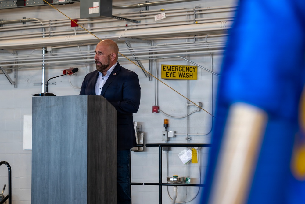 A bald man with a beard, dressed in a dark navy suit, is speaking at a gray podium equipped with a microphone. The setting appears to be an industrial or utility facility, evidenced by exposed piping, safety equipment signage, and a yellow 'Emergency Eye Wash' sign on the wall. A blue object, likely a flag or banner on a pole, is partially visible in the foreground right.