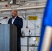 A bald man with a beard, dressed in a dark navy suit, is speaking at a gray podium equipped with a microphone. The setting appears to be an industrial or utility facility, evidenced by exposed piping, safety equipment signage, and a yellow 'Emergency Eye Wash' sign on the wall. A blue object, likely a flag or banner on a pole, is partially visible in the foreground right.
