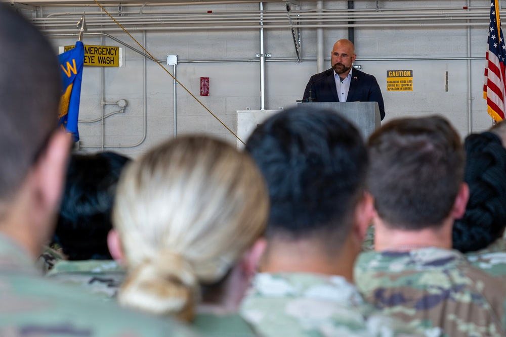 A civilian speaker addresses a military audience from a podium inside what appears to be a hangar facility. The audience consists of multiple uniformed military personnel seated with their backs to the camera. The setting suggests an official ceremony or briefing event at a military installation.