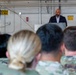 A civilian speaker addresses a military audience from a podium inside what appears to be a hangar facility. The audience consists of multiple uniformed military personnel seated with their backs to the camera. The setting suggests an official ceremony or briefing event at a military installation.