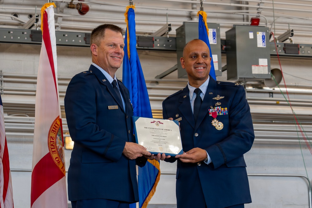Two U.S. Air Force officers in dress blue uniforms are jointly holding a certificate during a formal military ceremony. The certificate is visibly titled 'The Legion of Merit,' a U.S. military decoration. The event appears to take place inside a military hangar, with the Florida state flag and Air Force flags visible in the background.