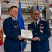 Two U.S. Air Force officers in dress blue uniforms are jointly holding a certificate during a formal military ceremony. The certificate is visibly titled 'The Legion of Merit,' a U.S. military decoration. The event appears to take place inside a military hangar, with the Florida state flag and Air Force flags visible in the background.