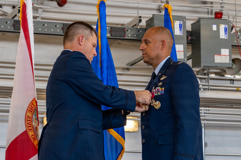 A military decoration ceremony is taking place in what appears to be a hangar facility. A man in a civilian dark suit is pinning a medal or award onto the uniform of a military officer wearing a US Air Force dress uniform adorned with multiple ribbons and badges. The Florida state flag and a blue military flag with gold fringe are visible in the background.