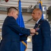 A military decoration ceremony is taking place in what appears to be a hangar facility. A man in a civilian dark suit is pinning a medal or award onto the uniform of a military officer wearing a US Air Force dress uniform adorned with multiple ribbons and badges. The Florida state flag and a blue military flag with gold fringe are visible in the background.