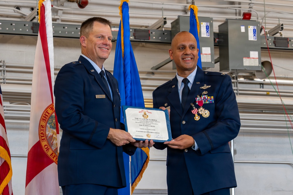 Two U.S. Air Force officers are posing together holding a certificate from the State of Florida titled 'The Florida Cross' for Extraordinary Meritorious Service. The ceremony appears to be taking place inside a military hangar, with the Florida state flag and blue Air Force flags visible in the background. The officer on the right is wearing multiple medals and decorations, suggesting this is a formal military awards ceremony.