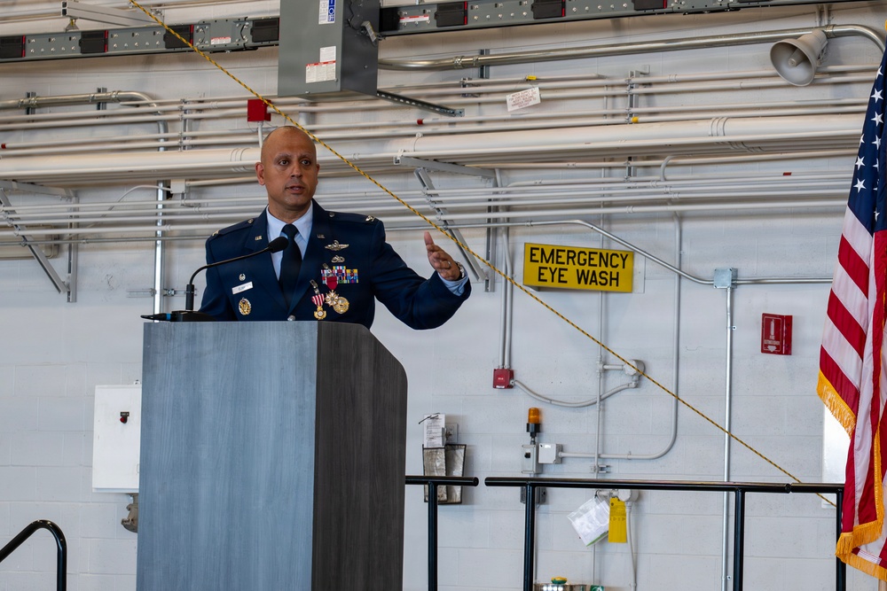 A U.S. Air Force officer in dress blue uniform is delivering a speech at a podium in what appears to be a maintenance hangar or industrial facility. The officer is wearing multiple medals and decorations, including what appear to be pilot wings, and is gesturing with one hand while speaking. An American flag is visible to the right of the frame.
