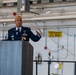 A U.S. Air Force officer in dress blue uniform is delivering a speech at a podium in what appears to be a maintenance hangar or industrial facility. The officer is wearing multiple medals and decorations, including what appear to be pilot wings, and is gesturing with one hand while speaking. An American flag is visible to the right of the frame.