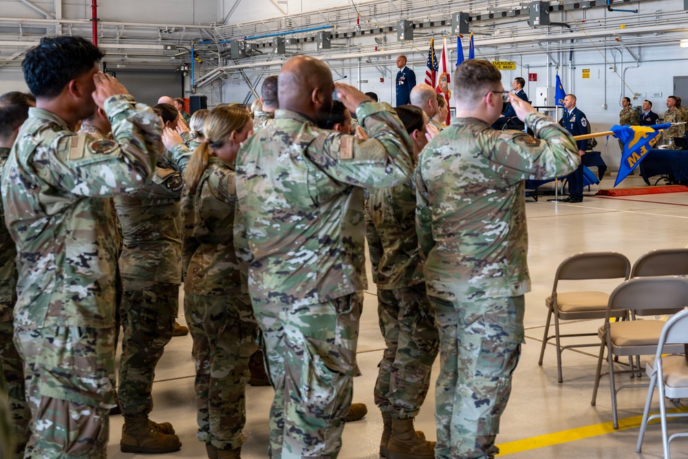 A group of military personnel in OCP (Operational Camouflage Pattern) uniforms are rendering a salute during what appears to be a formal military ceremony, likely a change of command or activation ceremony held inside a hangar facility. At the front of the room, officers in dress blue uniforms stand on a platform near flags including the American flag and what appears to be a unit guidon. The setting and attire suggest this is a U.S. Air Force or Space Force formal ceremony.