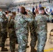 A group of military personnel in OCP (Operational Camouflage Pattern) uniforms are rendering a salute during what appears to be a formal military ceremony, likely a change of command or activation ceremony held inside a hangar facility. At the front of the room, officers in dress blue uniforms stand on a platform near flags including the American flag and what appears to be a unit guidon. The setting and attire suggest this is a U.S. Air Force or Space Force formal ceremony.