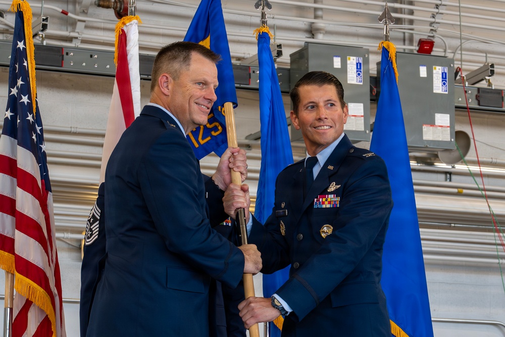 Two U.S. Air Force officers are participating in a formal guidon/flag passing ceremony, likely a change of command or assumption of command event. Both individuals are dressed in Air Force Service Dress uniforms and are jointly holding a wooden guidon staff. The ceremony is taking place inside what appears to be a military hangar, with the U.S. flag and blue Air Force flags visible in the background.