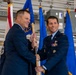 Two U.S. Air Force officers are participating in a formal guidon/flag passing ceremony, likely a change of command or assumption of command event. Both individuals are dressed in Air Force Service Dress uniforms and are jointly holding a wooden guidon staff. The ceremony is taking place inside what appears to be a military hangar, with the U.S. flag and blue Air Force flags visible in the background.