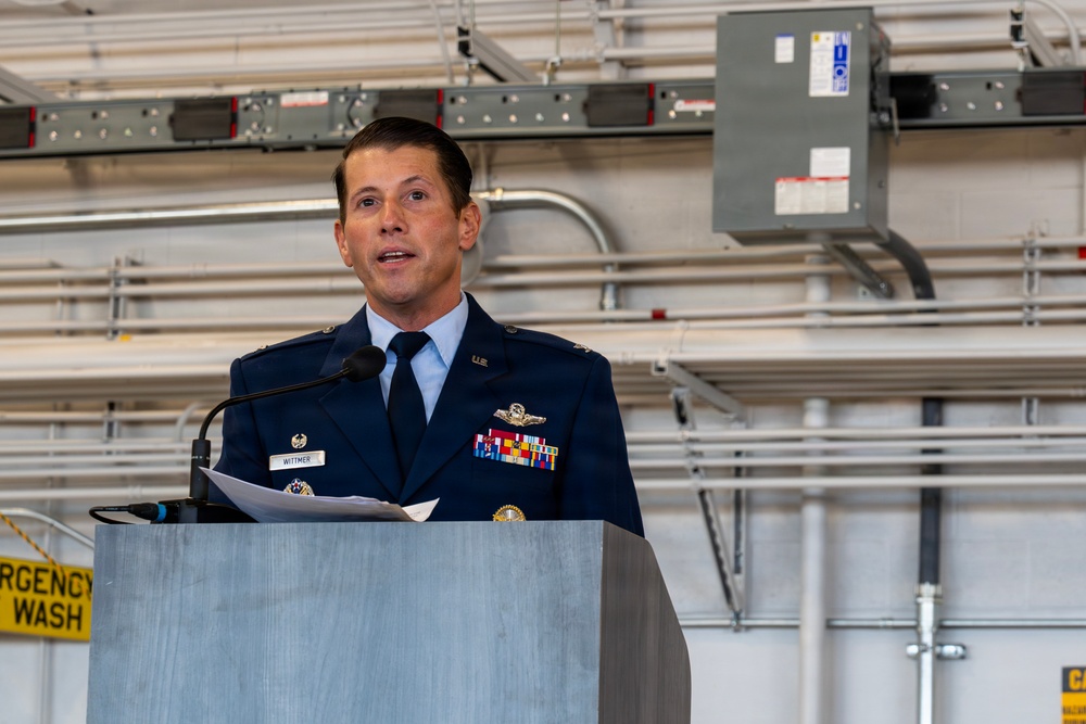 A U.S. Air Force officer in dress blues is speaking at a podium, holding papers and addressing an audience. His name tag reads 'WITTMER' and he is wearing multiple service ribbons, a command pilot badge, and other military decorations. The setting appears to be inside a military hangar or maintenance facility.