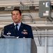 A U.S. Air Force officer in dress blues is speaking at a podium, holding papers and addressing an audience. His name tag reads 'WITTMER' and he is wearing multiple service ribbons, a command pilot badge, and other military decorations. The setting appears to be inside a military hangar or maintenance facility.