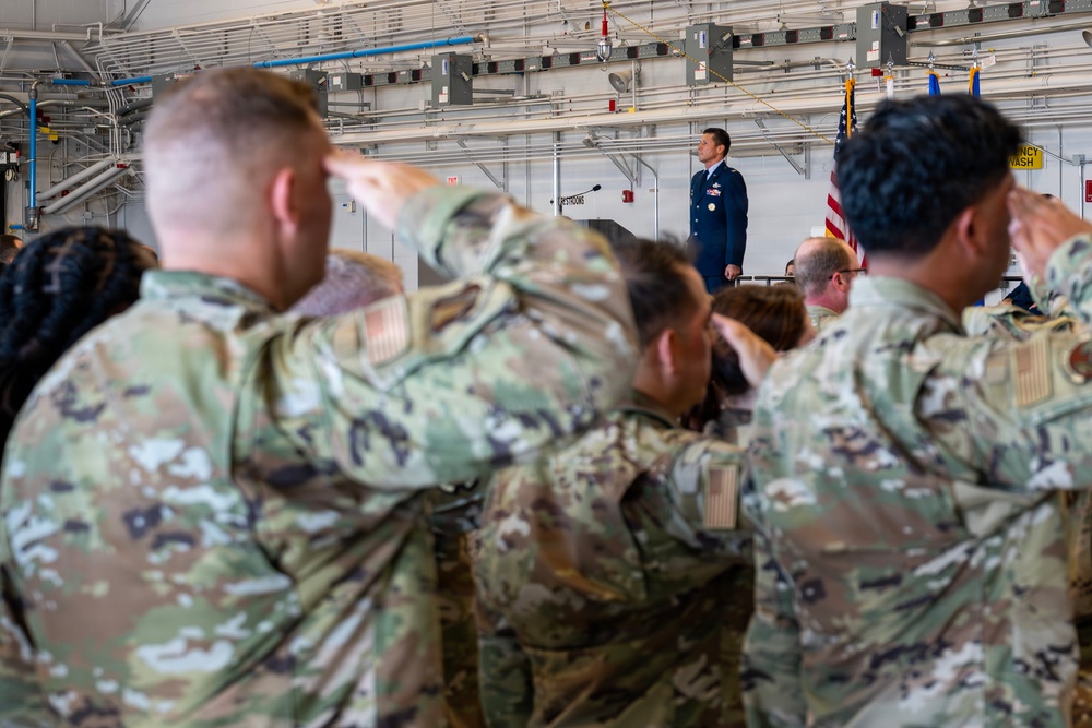 A military ceremony is taking place inside what appears to be a hangar or large indoor facility. Multiple service members in OCP (Operational Camouflage Pattern) uniforms are rendering a salute toward a single officer standing at a podium in dress blue uniform. An American flag and at least one additional flag are visible near the podium.