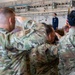 A military ceremony is taking place inside what appears to be a hangar or large indoor facility. Multiple service members in OCP (Operational Camouflage Pattern) uniforms are rendering a salute toward a single officer standing at a podium in dress blue uniform. An American flag and at least one additional flag are visible near the podium.