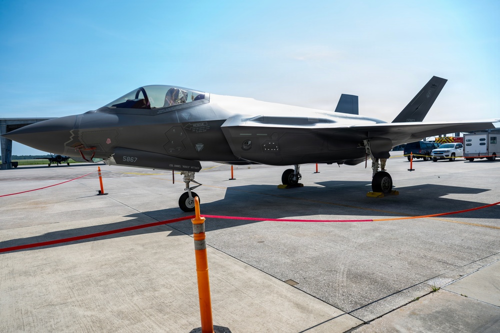 A Lockheed Martin F-35 Lightning II stealth fighter jet is displayed on a tarmac, likely at an airshow or static display event. The aircraft bears the serial number 5867 and is cordoned off with orange safety cones and a red rope barrier. Support vehicles and ground equipment are visible in the background.