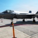 A Lockheed Martin F-35 Lightning II stealth fighter jet is displayed on a tarmac, likely at an airshow or static display event. The aircraft bears the serial number 5867 and is cordoned off with orange safety cones and a red rope barrier. Support vehicles and ground equipment are visible in the background.