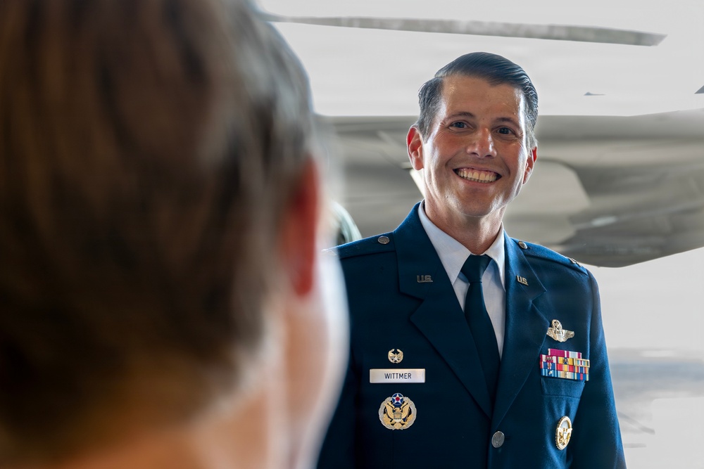 A U.S. Air Force officer in dress blues uniform is smiling broadly, appearing to be engaged in conversation with another person whose back is visible in the foreground. The officer's name tag reads 'WITTMER' and he is wearing multiple service ribbons, pilot wings, and other military insignia. The setting appears to be inside or near an aircraft hangar with a large aircraft visible in the background.