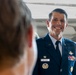 A U.S. Air Force officer in dress blues uniform is smiling broadly, appearing to be engaged in conversation with another person whose back is visible in the foreground. The officer's name tag reads 'WITTMER' and he is wearing multiple service ribbons, pilot wings, and other military insignia. The setting appears to be inside or near an aircraft hangar with a large aircraft visible in the background.