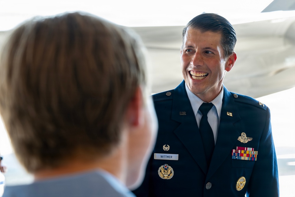 A U.S. Air Force officer named Wittmer, wearing a dress blue uniform with multiple service ribbons and badges, is smiling broadly during what appears to be a formal military event or ceremony. He is engaged in conversation with another individual whose back is to the camera. The setting appears to be inside or near an aircraft hangar, with part of an aircraft visible in the background.