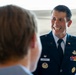 A U.S. Air Force officer named Wittmer, wearing a dress blue uniform with multiple service ribbons and badges, is smiling broadly during what appears to be a formal military event or ceremony. He is engaged in conversation with another individual whose back is to the camera. The setting appears to be inside or near an aircraft hangar, with part of an aircraft visible in the background.