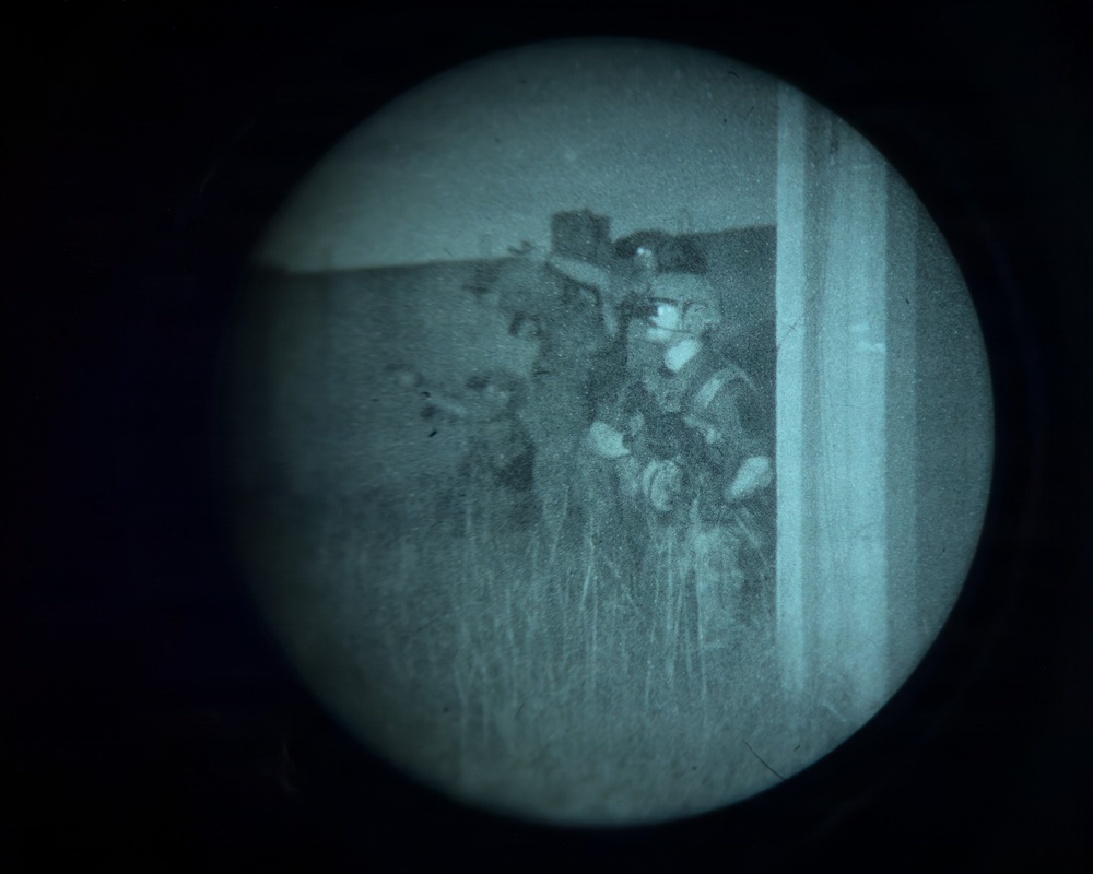 A night vision device (NVD) captured image showing one or more individuals in tactical gear positioned near a structure or doorway. The circular vignette is characteristic of looking through a night vision monocular or binocular optic. The scene appears to depict personnel in a field or grassy environment at night.