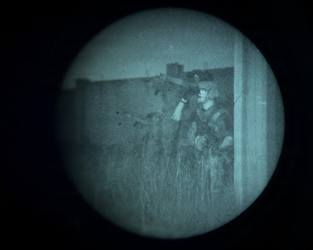 A night-vision or image-intensifier optic view showing one or more figures in tactical/military gear positioned near a structure. The circular vignette and teal-green tonal rendering are characteristic of generation-2 or generation-3 night-vision devices. The figures appear to be in a field or overgrown area adjacent to a building or wall.