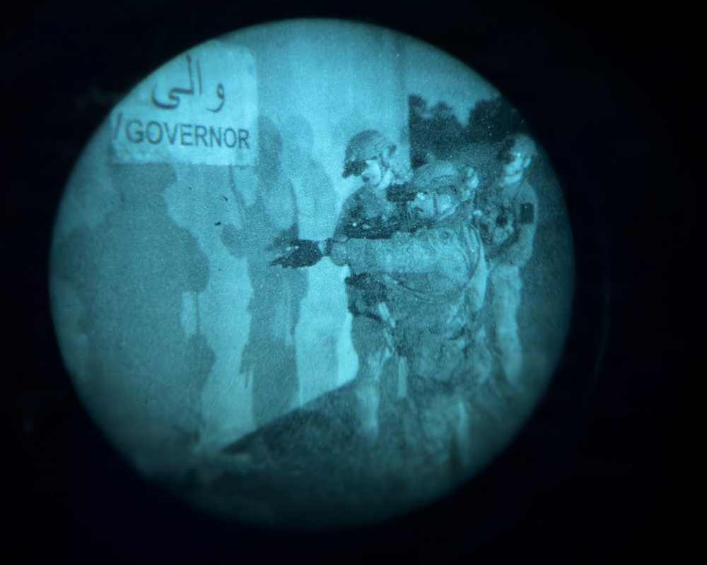 Image captured through a night-vision device showing armed military personnel in full combat gear positioned near a building entrance. A bilingual sign reading 'GOVERNOR' in English and Arabic script (والي) is visible on the wall. The circular vignette and teal-green tonal quality are characteristic of night-vision optics.