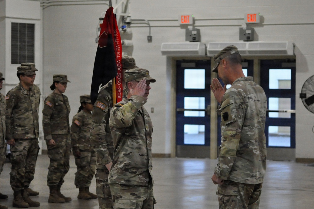 A military ceremony is taking place inside an indoor facility, likely a drill hall or armory. Two soldiers in the foreground are exchanging salutes, with one holding a unit guidon/flag. Additional soldiers stand in formation in the background.