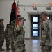 A military ceremony is taking place inside an indoor facility, likely a drill hall or armory. Two soldiers in the foreground are exchanging salutes, with one holding a unit guidon/flag. Additional soldiers stand in formation in the background.