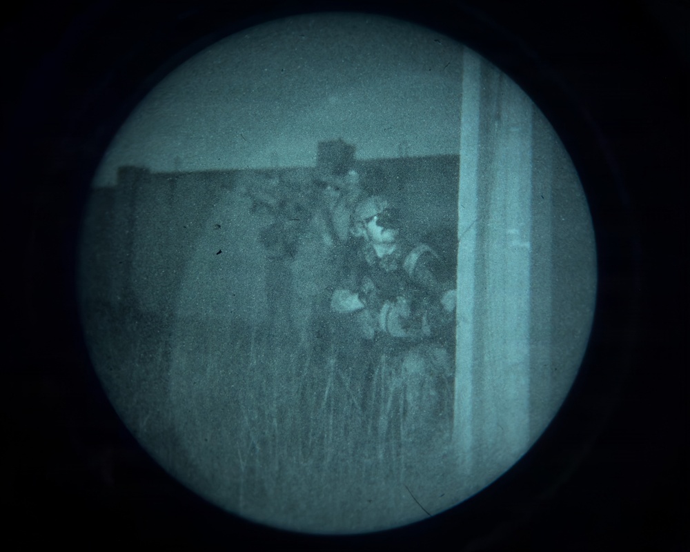 A night vision device (NVD) captured image showing what appears to be multiple armed personnel in tactical gear positioned near a structure or wall. The circular field of view is characteristic of a night vision monocular or scope. The green-tinted monochromatic image shows figures in a low-light outdoor environment with tall grass visible in the foreground.